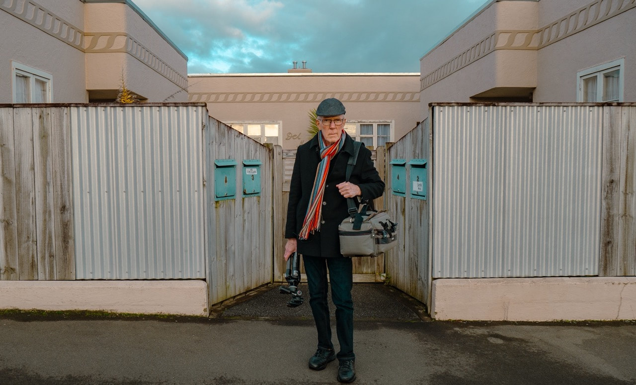 A photograph of photographer Richard Wotton in the middle of the picture looking straight into the camera. He is dressed in a black overcoat, striped red and blue scarf, blue jeans, a cloth cap hat, and dark trainers. He is carrying a tripod in his right hand and has a camera bag on his left shoulder. Behind him are the pale brown and yellow cement clad buildings of Del Mer Flats, partially hidden behind a tall wooden slat fence. Richard is standing in front of the entrance path where the fence diverts inwards, the with each side of the fence supporting two pale blue mailboxes. The sky is overcast with blue patches. The photo was taken in winter 2022 during the production of Richard Wotton: Moments Later. Credit Kevin Double. License CC BY NC 2.0