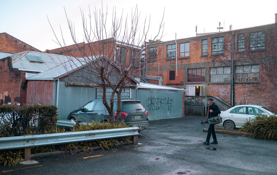 A photograph of the rear of brick buildings in a back alley off of Watt Street in Whanganui. Walking into the frame is photographer Richard Wotton. He is looking up towards the buildings, carrying a tripod and camera bag. He is wearing a black overcoat, blue jeans, and a red and blue striped scarf, plus black trainers. The weather is bright and overcast with recent rain evident. It is in the middle of winter time. Credit Kevin Double. Taken during the 2022 production of Richard Wotton: Moments Later in Watt Street, Whanganui, New Zealand.