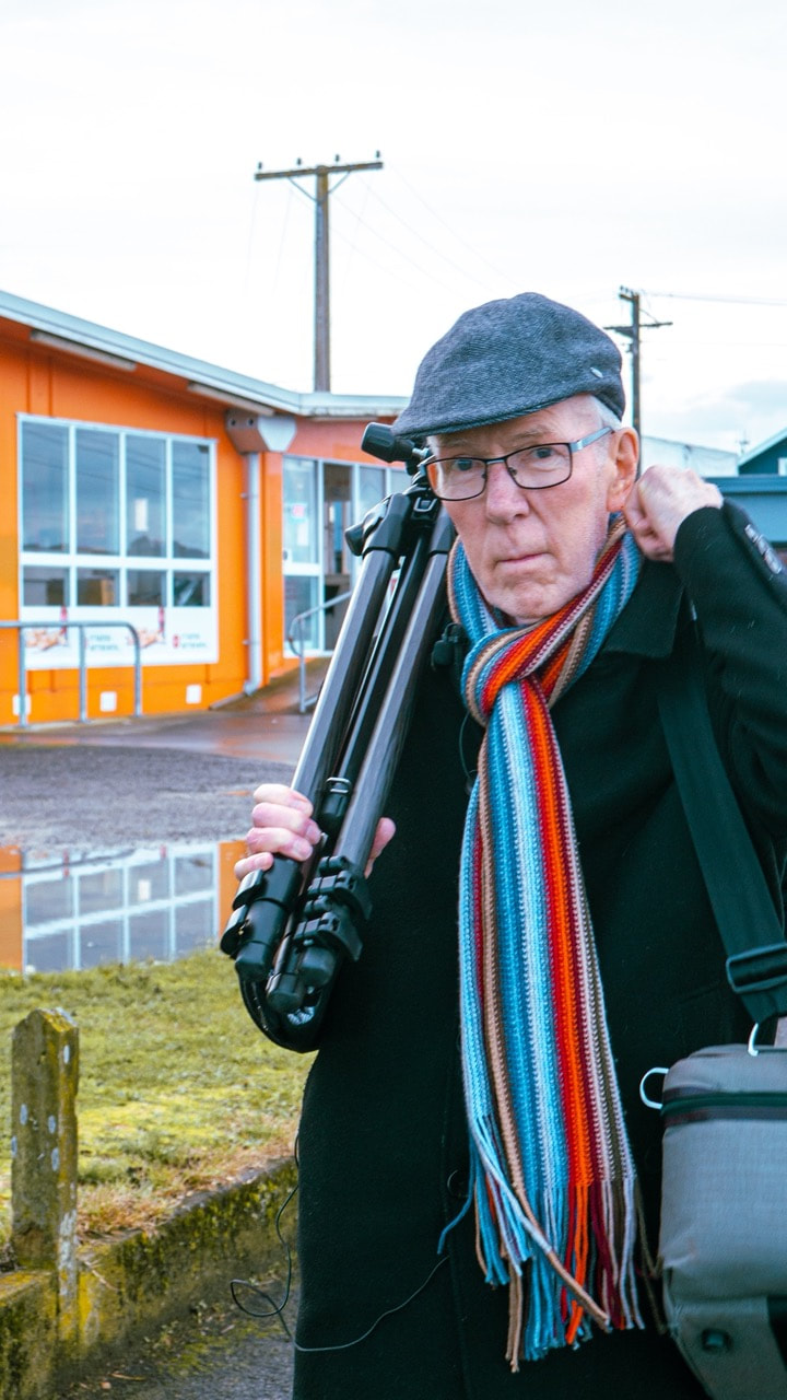 A photograph of the photographer Richard Wotton in his mid 70s looking at the camera. He is wearing a heavy black overcoat, red and blue striped woollen scarf, a peaked cloth hat, and glasses. He is carrying a tripod on his right shoulder and a camera bag on his left. He is looking directly into the camera. Behind him is the orange building known as Somme Parade Takeaways. The photo was taken in 2022 during the filming of RICHARD WOTTON: MOMENTS LATER. Credit Kevin Double. Location Somme Parade, Whanganui.
